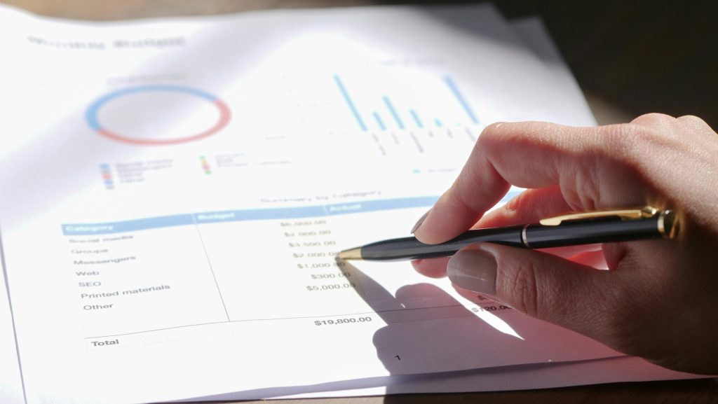 Hand pointing at engineering budget on printed cost breakdown sheet with charts in background